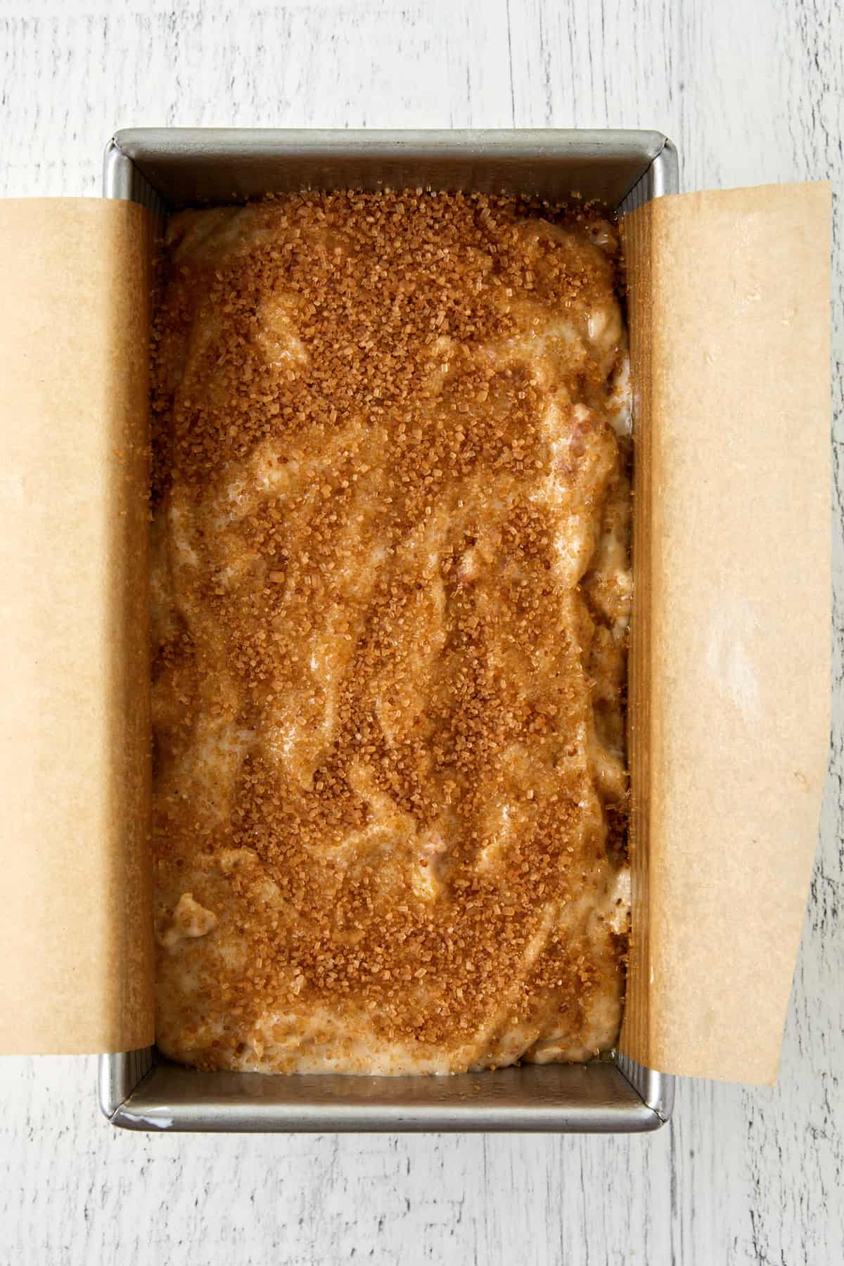 Overhead view of banana rhubarb bread batter in a loaf pan with turbinado sugar on top and lined with parchment paper.