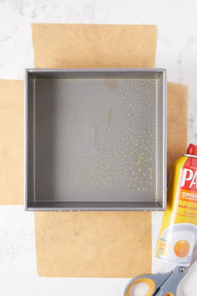 The image shows an empty metal baking pan placed on top of parchment paper, which is on a marble countertop. The parchment paper beneath it appears to be cut to fit the pan, possibly in preparation for baking. The perspective is from above, providing a clear view of the pan and the parchment setup.