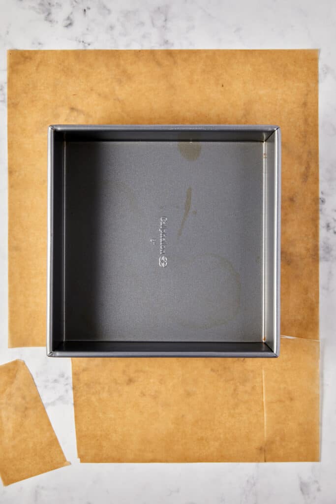 The image shows an empty metal baking pan placed on top of parchment paper, which is on a marble countertop.  The parchment paper beneath it appears to be cut to fit the pan, possibly in preparation for baking. The perspective is from above, providing a clear view of the pan and the parchment setup.