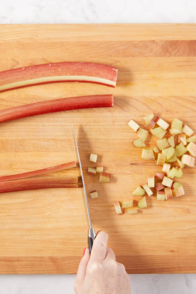 overhead view of rhubarb stalks on a cutting board being cut with a knife into cubes.