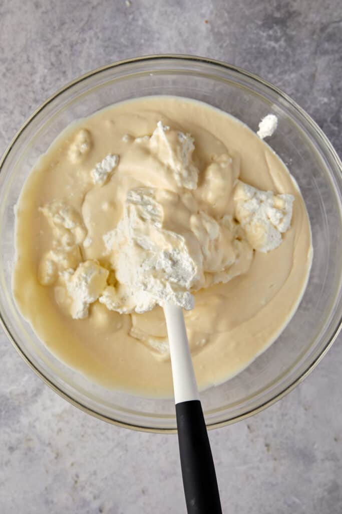 Overhead view of folding whipped cream in banana pudding in a glass bowl.
