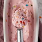 A close-up of a metal ice cream scoop lifting a scoop of pink ice cream with fruity pebbles cereal pieces from a white container.