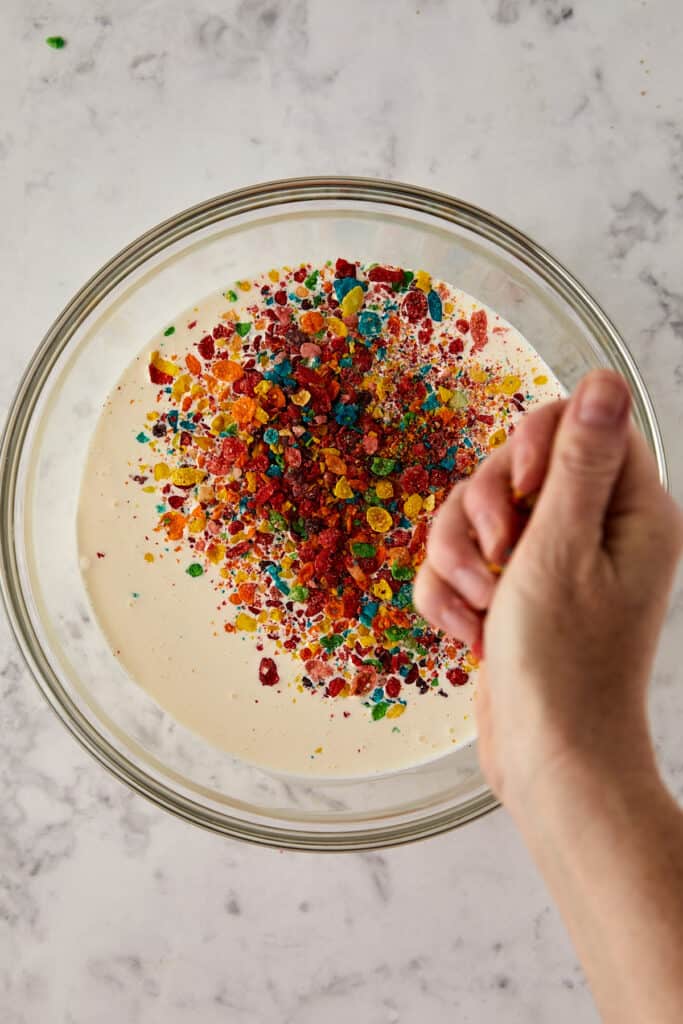 Overhead view of fruity pebbles being crushed by a hand over the glass bowl into the heavy cream and milk. 