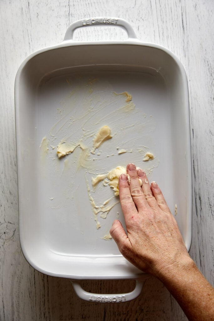 Overhead view of casserole dish with butter being smeared on the bottom with a hand.