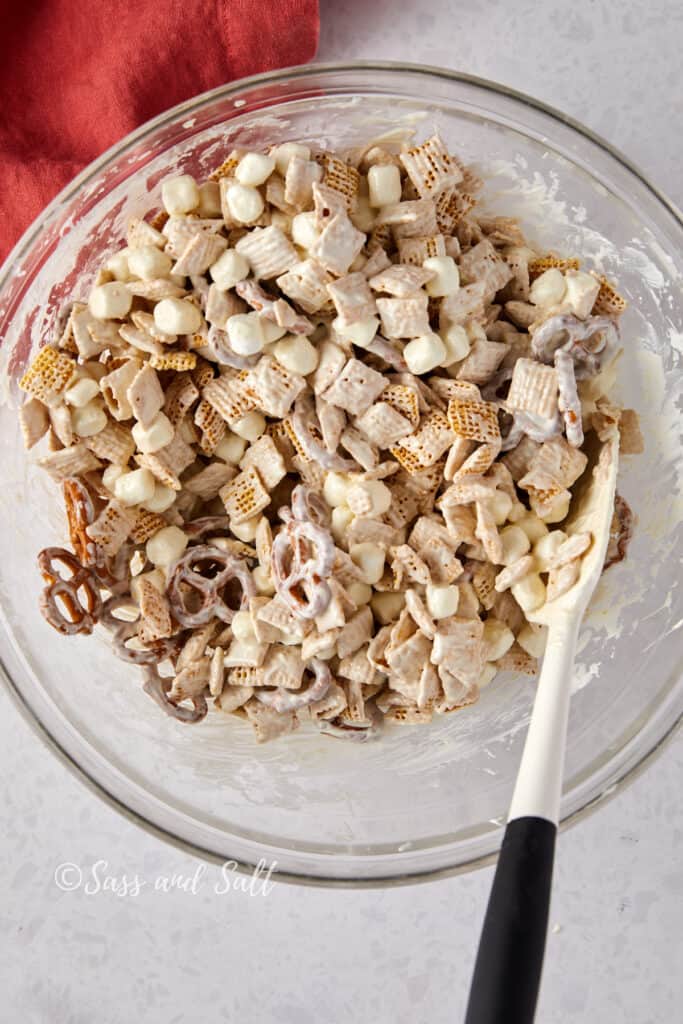 Overhead view of white chocolate mixed with Chex mix with a spatula in a glass bowl.
