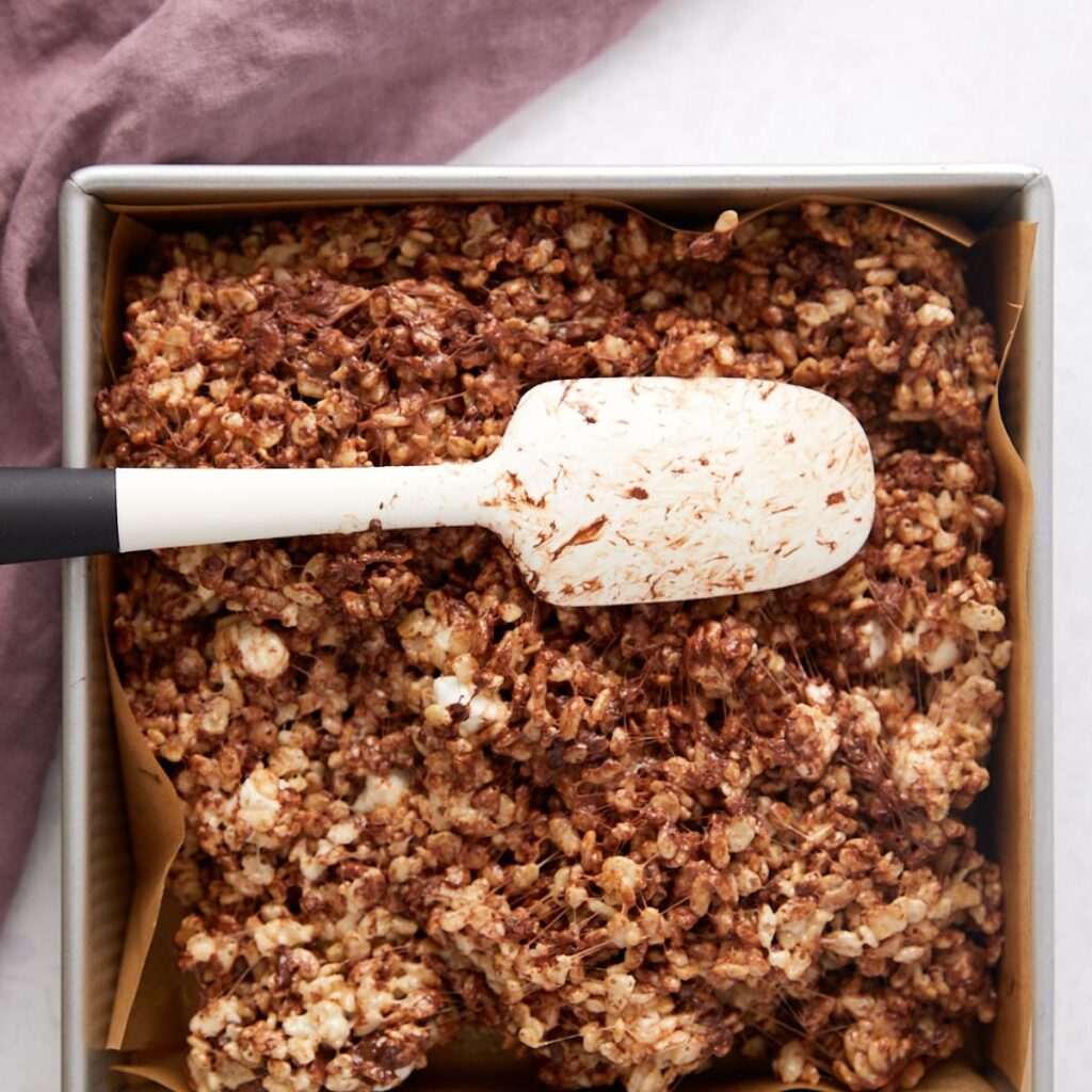 A baking pan lined with parchment paper holds delicious Chocolate Chip Rice Krispie Treats. A white and black spatula with chocolate residue rests on top, while a purple cloth is partially visible beside the pan.