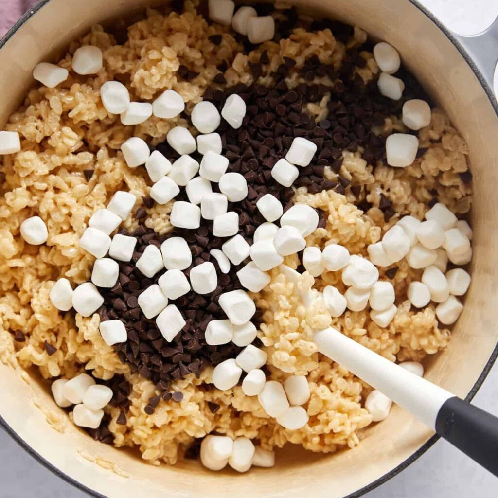 A pot of rice krispie treat mixture is being stirred with a spatula. Mini marshmallows and chocolate chips are scattered on top, making this the perfect Chocolate Chip Rice Krispie Treats.
