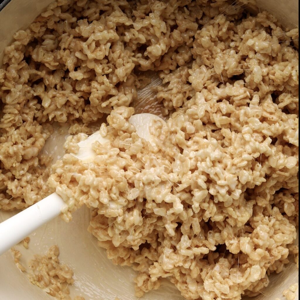 Close-up of a white spatula mixing Rice Krispies cereal with melted marshmallows and chocolate chips in a large bowl, creating a sticky, creamy mixture for delicious rice krispie treats with chocolate chips.