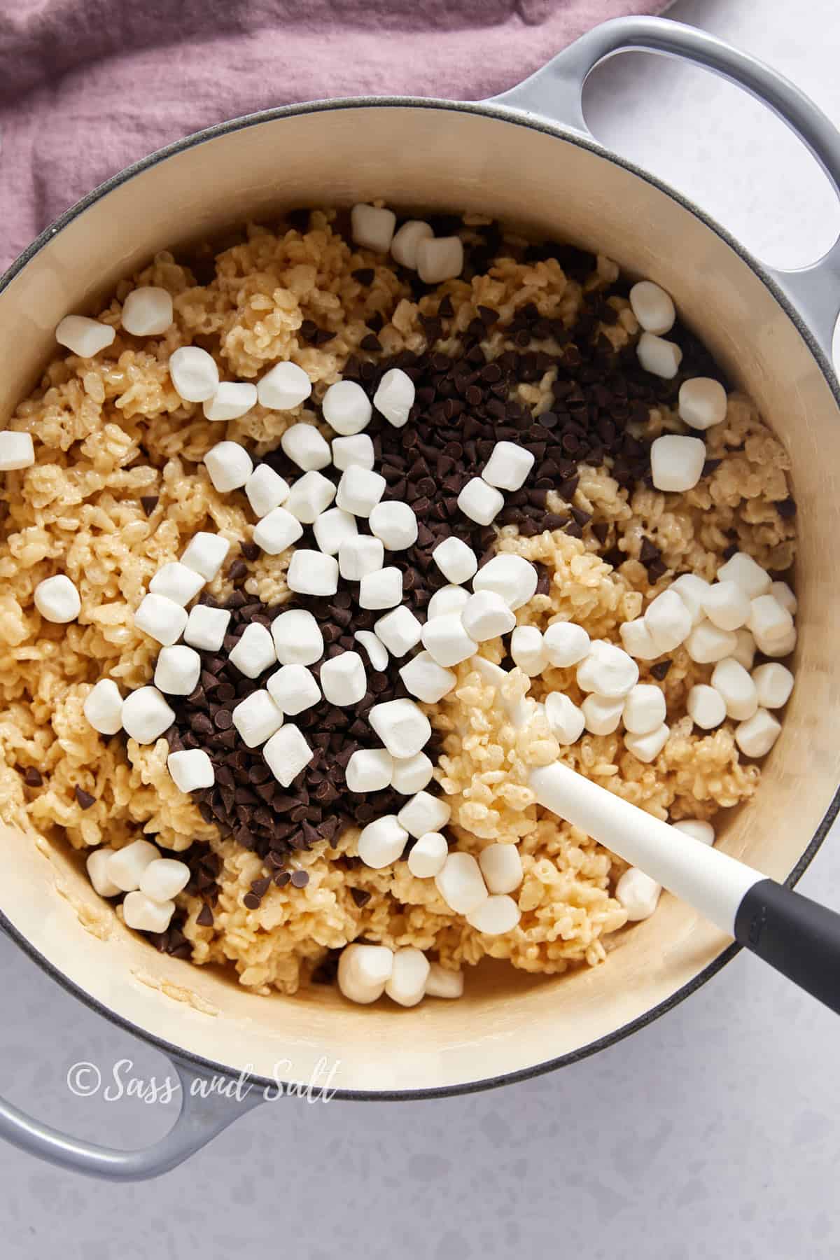 Overhead view of mini chocolate chips and mini marshmallows on top of rice krispies and melted marshmallows in dutch oven.