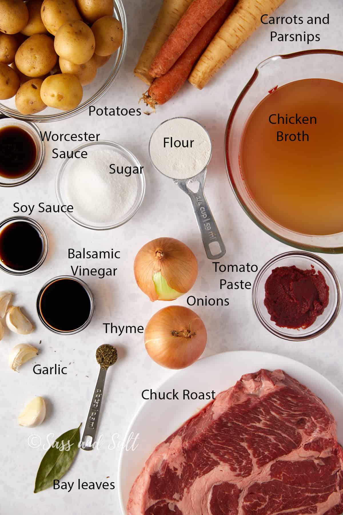 This photo depicts an array of ingredients laid out on a light surface, each labeled for a beef stew recipe. From the top left corner, there is a bowl of small potatoes, followed by carrots and parsnips to their right. Below the vegetables, there is chicken broth in a glass measuring cup. To the left of the broth, there are small dishes containing flour and sugar, and below them, Worcester sauce and soy sauce. Moving to the bottom left of the image, garlic cloves and bay leaves sit beside a spoonful of dried thyme. In the center, balsamic vinegar is in a small dish, with two onions to its right and a can of tomato paste above. Dominating the bottom right of the image is a large piece of chuck roast on a white plate. The photo is watermarked with "Sass and Salt."