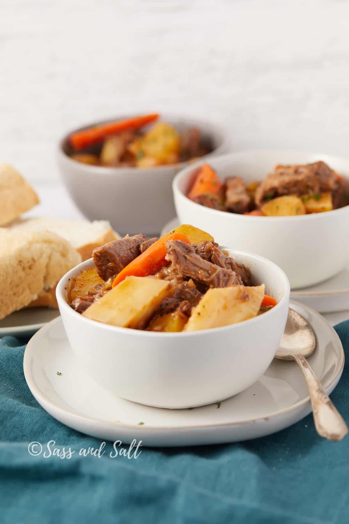 The image features a styled presentation of beef stew served in two white bowls. The stew is garnished with chopped herbs and is accompanied by pieces of rustic bread on a textured white table. The bowls are placed on a teal cloth, and a silver spoon rests beside one of the bowls, ready for eating. The stew looks hearty with chunks of beef, carrots, and potatoes, and is captured in a well-lit setting that accentuates its colors and textures. The watermark "Sass and Salt" indicates the branding behind the image.