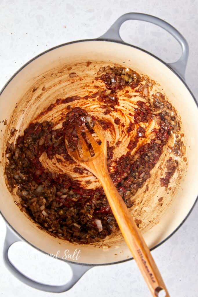 This photo captures an overhead view of a light-colored Dutch oven where onions have been sautéed, and soy sauce, Worcester sauce, and tomato paste have been added. The tomato paste is streaking across the bottom of the pot, showing the mixing process with a wooden spoon that is being used to combine the ingredients. The rich, red hue of the tomato paste contrasts with the light interior of the pot, indicating the start of layering flavors for a stew or sauce. The image is marked with "Sass and Salt."