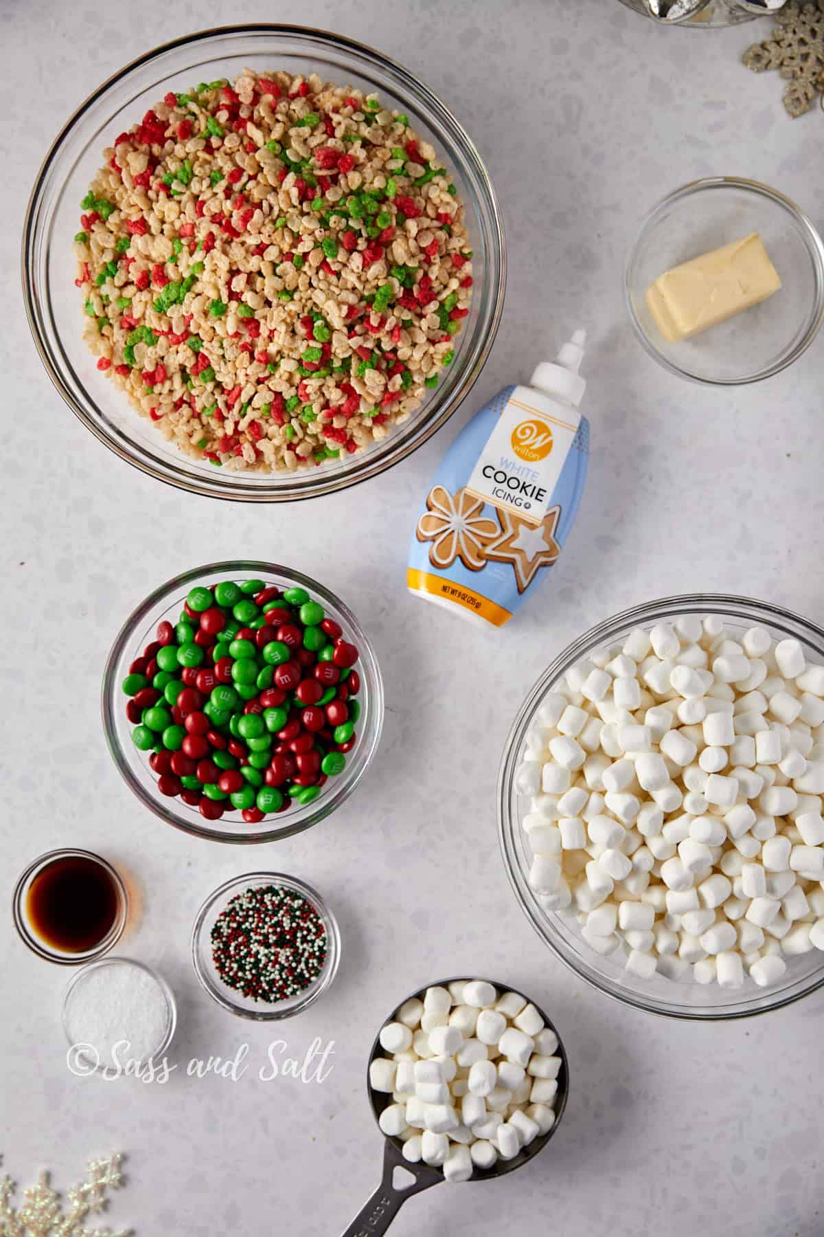 Overhead view of a kitchen counter with ingredients for Easy Rice Krispie Treats, including a bowl of Rice Krispies cereal, marshmallows, butter, vanilla extract, salt, sprinkles, frosting, and a colorful assortment of red and green M&M's, all neatly arranged and ready for preparation.