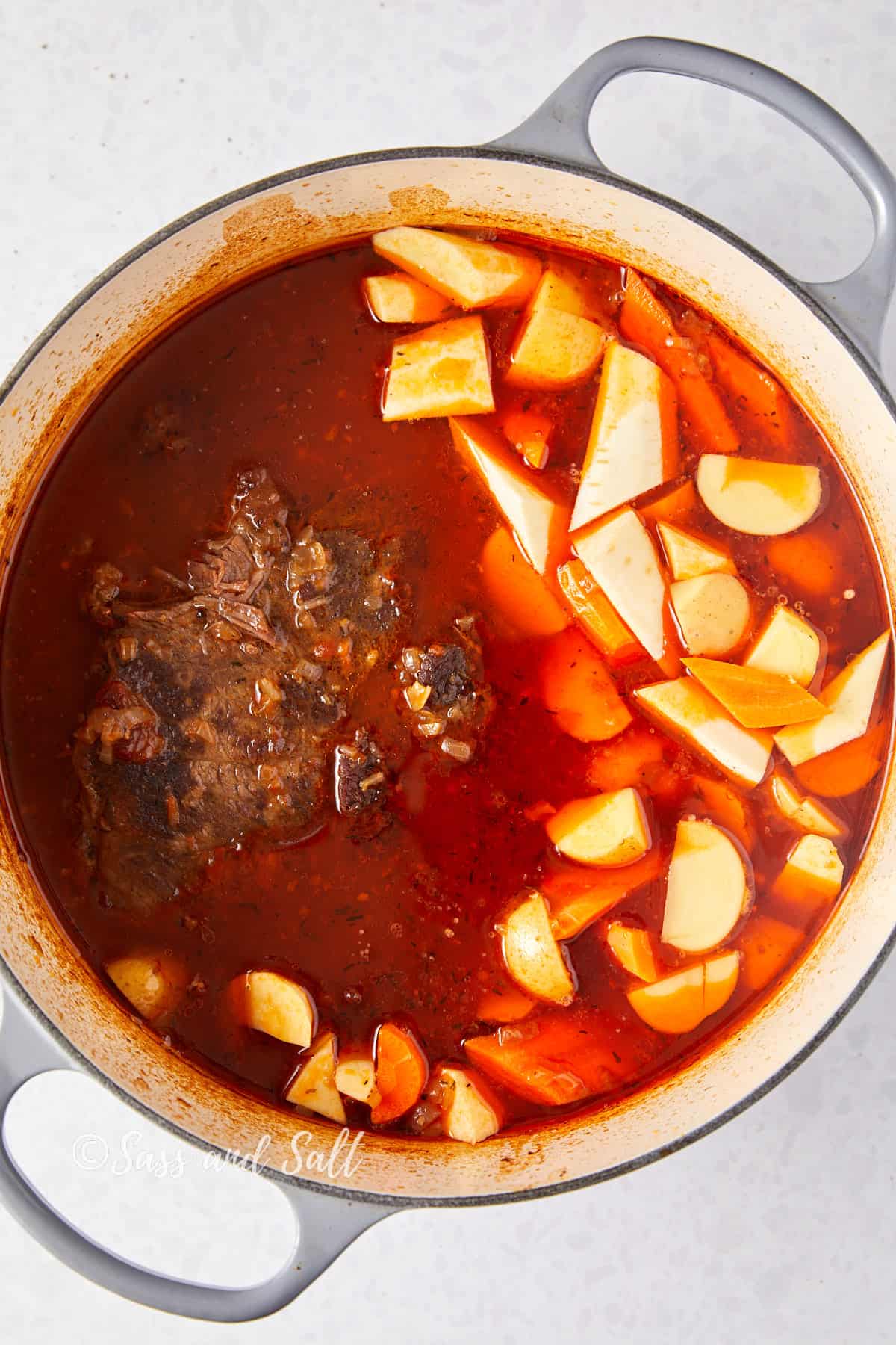 The image is an overhead view of a Dutch oven containing a hearty beef stew. The stew features a large piece of browned chuck roast surrounded by a rich, reddish-brown broth. Added to the stew are chunks of carrots and quartered potatoes, which are starting to cook in the flavorful liquid. The colors are vibrant, with the orange of the carrots and the cream of the potatoes standing out against the deep shade of the broth. The Dutch oven is on a light, speckled background, and "Sass and Salt" is watermarked onto the image.