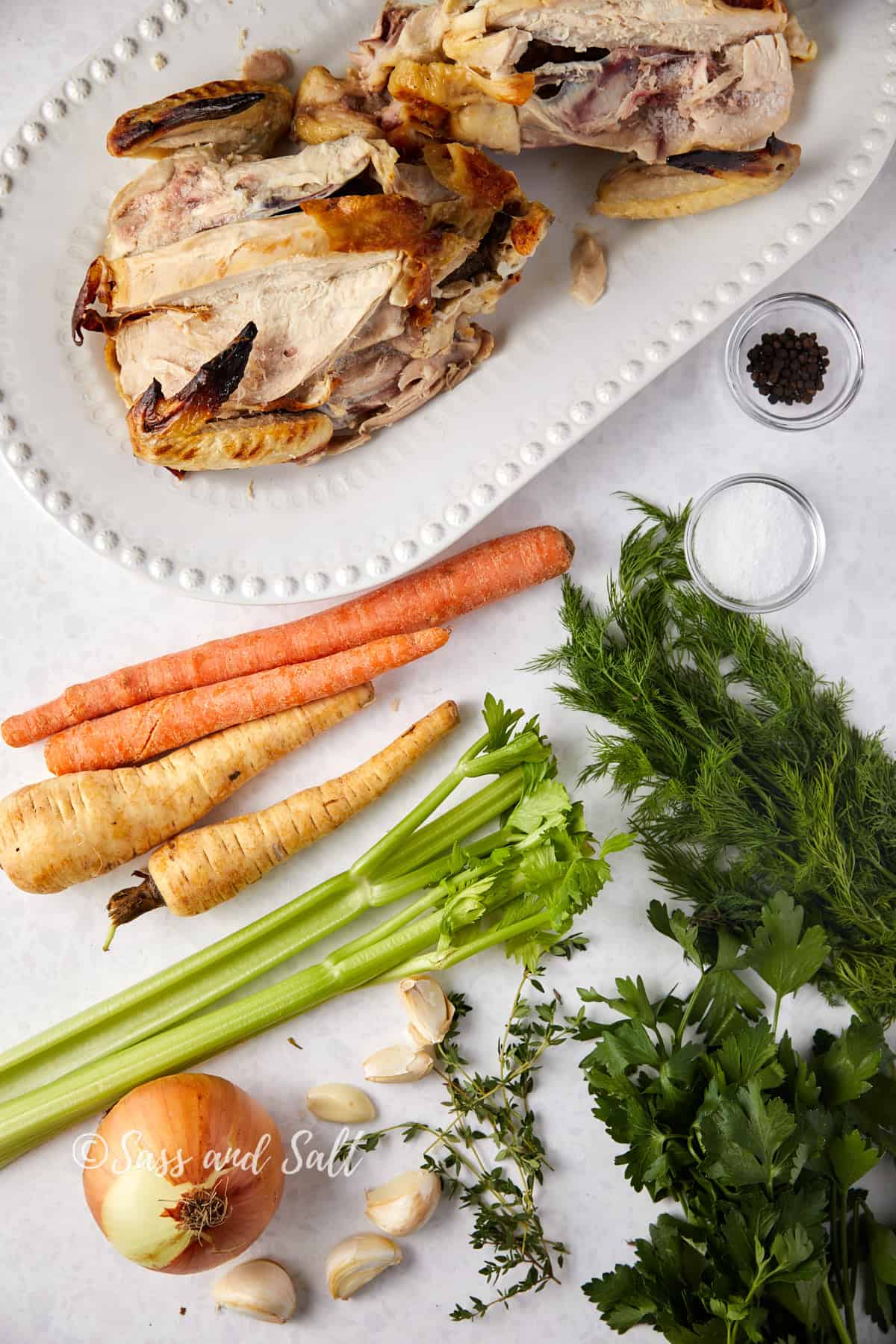 Overhead view of ingredients laid out on a white surface for making rotisserie chicken bone broth, including a white plate with leftover pieces of a rotisserie chicken, whole carrots, a bunch of celery, a large onion, several cloves of garlic, fresh herbs such as dill and parsley, and small bowls of salt and black peppercorns. The text "Sass and Salt" is overlaid at the bottom of the image.