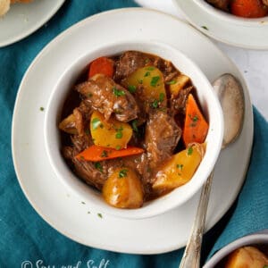 The image shows two bowls of hearty beef stew, garnished with chopped parsley. The stew contains chunks of beef, carrots, and potatoes, and is presented on a white marble surface with a slice of crusty bread on the side. Underneath the bowls is a teal cloth napkin, adding a pop of color to the setting. The photo is watermarked with "Sass and Salt."
