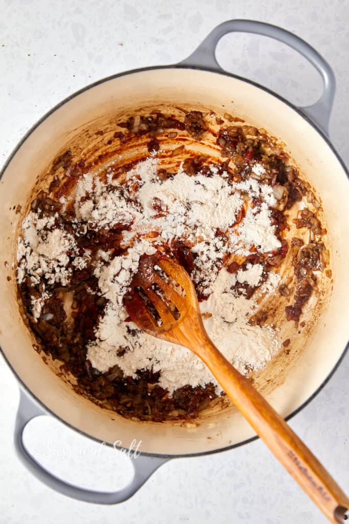 This photo is an overhead shot of a cream-colored Dutch oven with caramelized onions and remnants of cooked down ingredients at the bottom. Flour has been sprinkled over the mixture, ready to be stirred in as a thickener for a stew or sauce. A wooden spoon, which has been used to mix the ingredients, rests in the pot, indicating the cooking process is underway. The pot handles are visible, and the kitchen surface beneath is lightly speckled. The image is watermarked with "Sass and Salt."