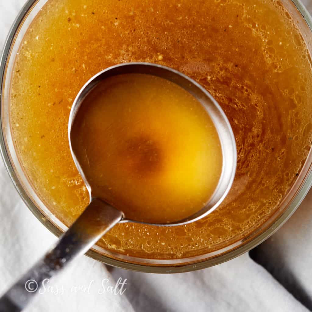 Overhead view of a clear glass bowl filled with golden rotisserie chicken bone broth, with a ladle partially submerged, showcasing the rich, translucent color of the broth. The bowl rests on a light, textured cloth.
