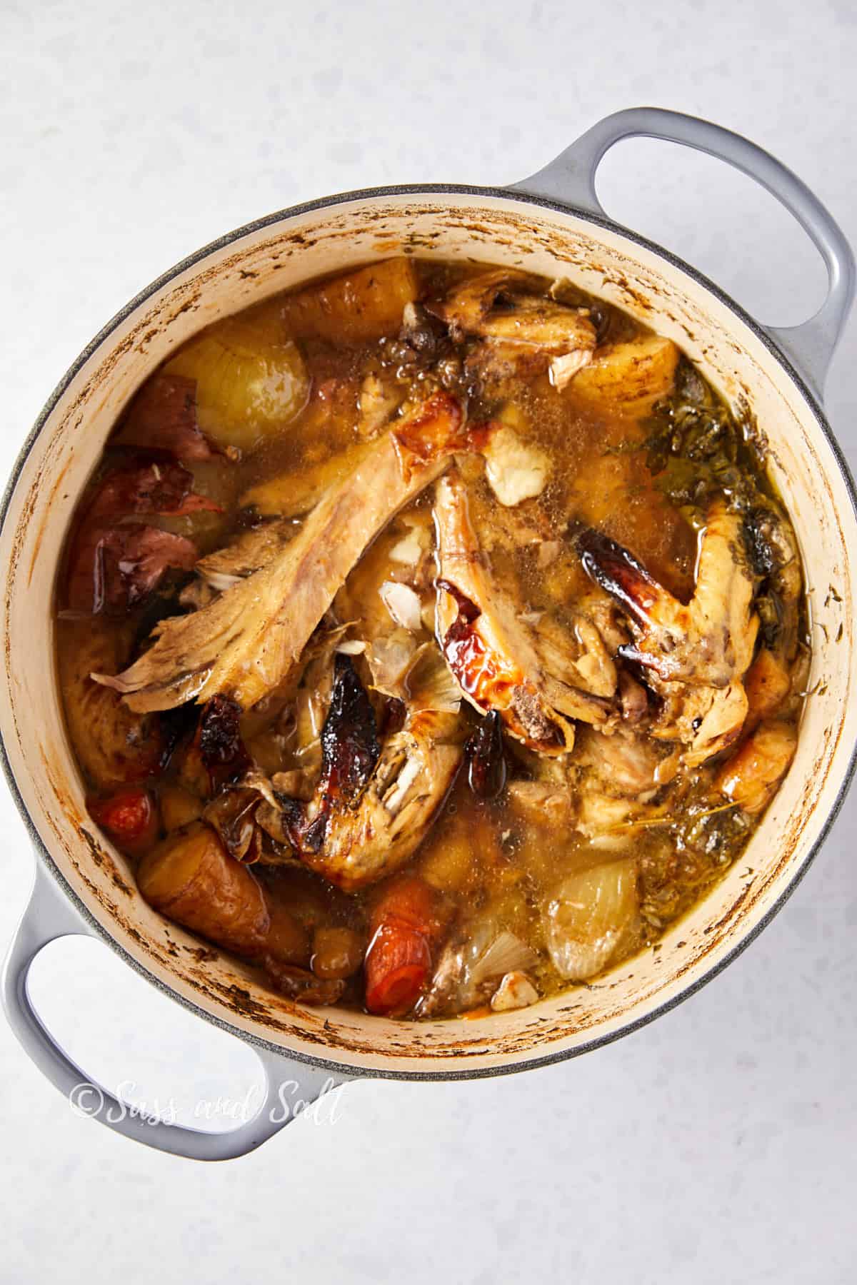 Overhead view of a pot filled with simmered rotisserie chicken bone broth, showing chicken bones with some meat attached, chunks of vegetables, and herbs in a rich, savory liquid. The sides of the pot have a slight browning from the cooking process, and the text "Sass and Salt" is marked at the bottom.





