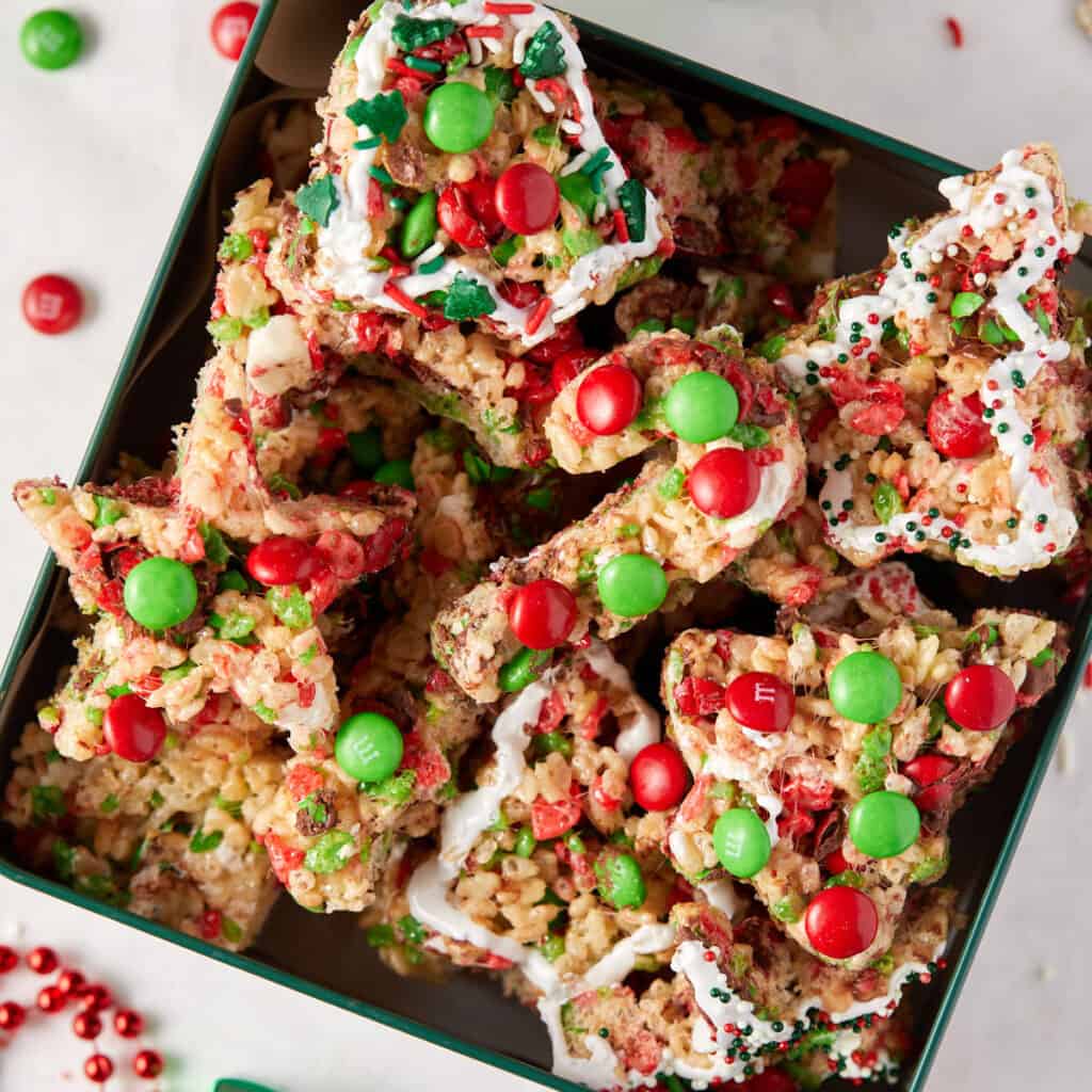 Overhead view of a Christmas tin filled with homemade Rice Krispie Treats, each generously drizzled with white frosting and adorned with colorful sprinkles, creating a festive and inviting holiday dessert.
