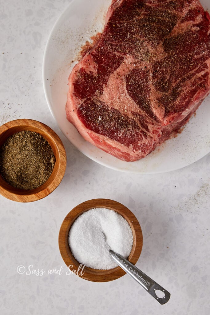 This image shows a raw chuck roast on a white plate, generously seasoned with black pepper and salt, ready for cooking. Above the plate, the seasonings are displayed in two small wooden bowls - one with ground black pepper and the other with white kosher salt, accompanied by a metal measuring spoon. The surface underneath is lightly speckled, suggesting a clean, minimalist preparation area. The watermark "Sass and Salt."