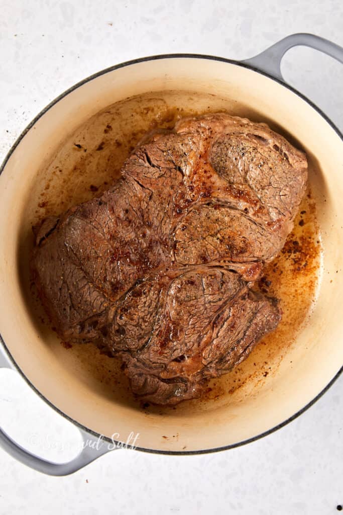 This picture shows a large piece of chuck roast browned to a golden color in a cream-colored Dutch oven. The sear on the beef has created a rich, caramelized crust, and there are bits of seasoning visible on its surface. Some juices have been released, contributing to the fond at the bottom of the pot, which will add depth to the stew's flavor. The Dutch oven handle is visible to the top right, indicating it's ready for the next step in the cooking process. The image is watermarked with "Sass and Salt."