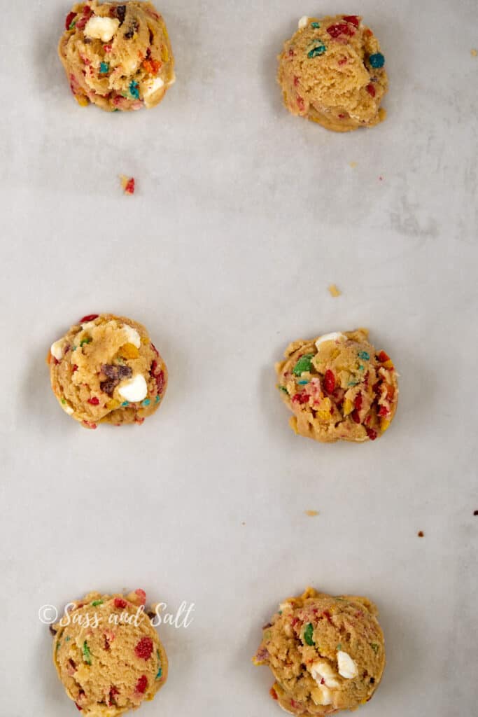 The image displays a baking sheet lined with parchment paper on which balls of cookie dough are arranged. The dough is dotted with colorful pieces of Fruity Pebbles cereal and white marshmallow bits, ready to be baked. The backdrop is clean and simple, focusing all attention on the vibrant dough portions.