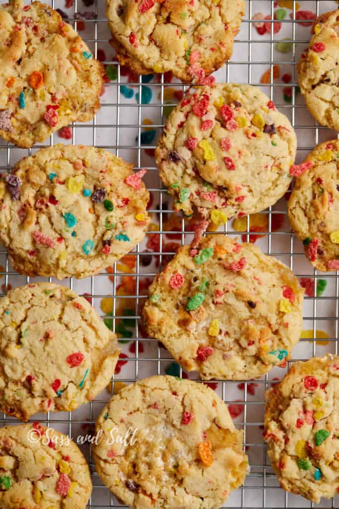 The image shows freshly baked cookies cooling on a wire rack. These treats are packed with colorful Fruity Pebbles cereal pieces that add a vibrant contrast to the golden-brown hue of the cookies.