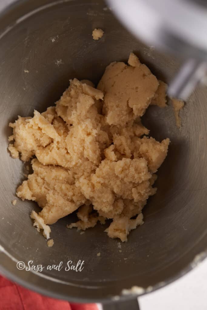 The image depicts a stainless steel mixing bowl where butter and a combination of brown and granulated sugars have been creamed together. The mixture appears uniformly blended, light in color, and fluffy in texture, with some clinging to the sides of the bowl, which indicates the ingredients are well mixed. The mixer's paddle attachment is in motion, suggesting the creaming process is ongoing.
