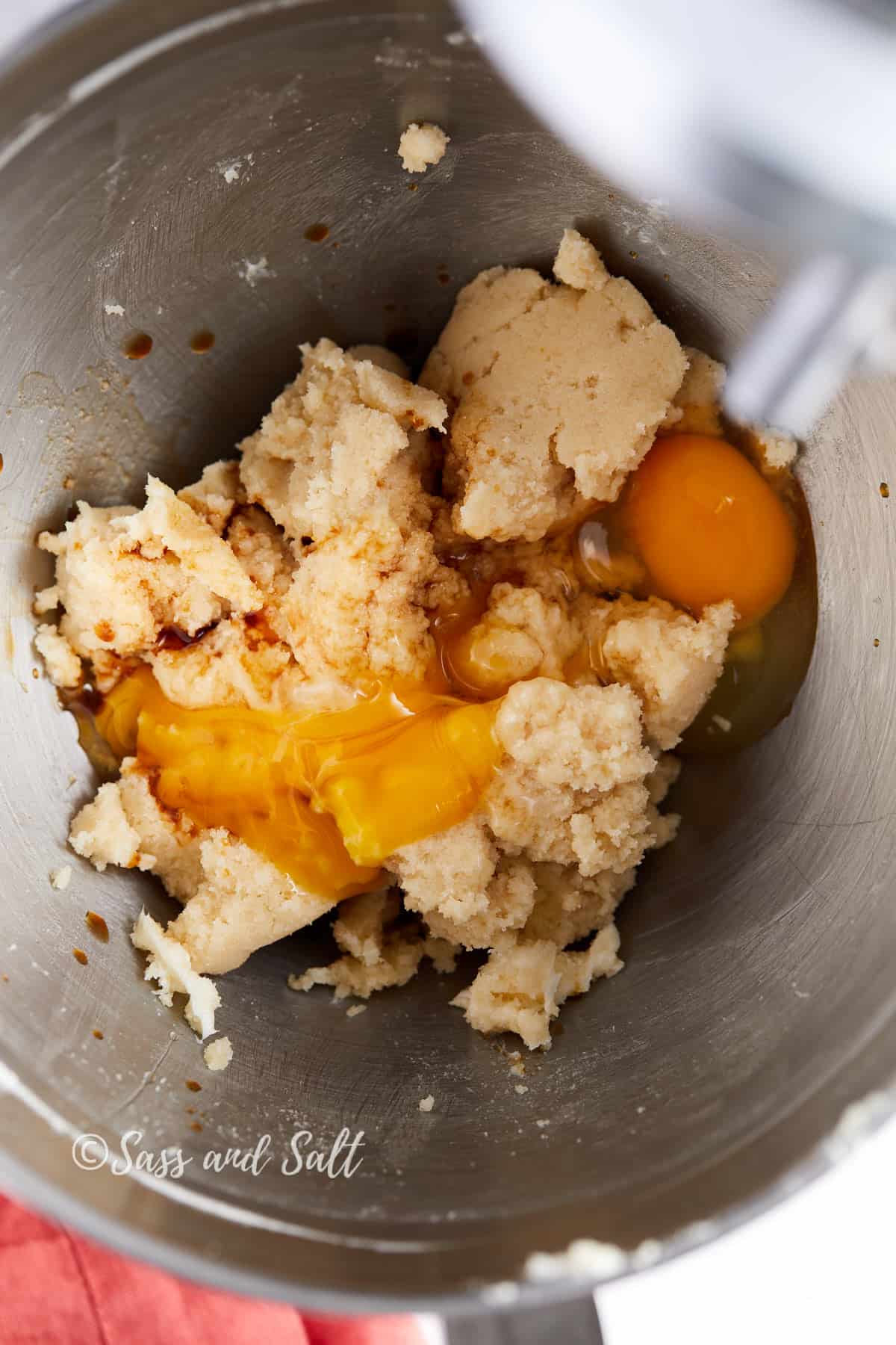 The image shows a close-up view of a mixing bowl where creamed butter and sugar mixture has been combined with eggs. Two whole eggs rest on top of the mixture, with the vibrant orange yolks standing out against the pale background of the dough. The process of adding wet ingredients to the creamed mixture is a crucial step in cookie-making, and the image captures this moment just before the eggs are mixed in.
