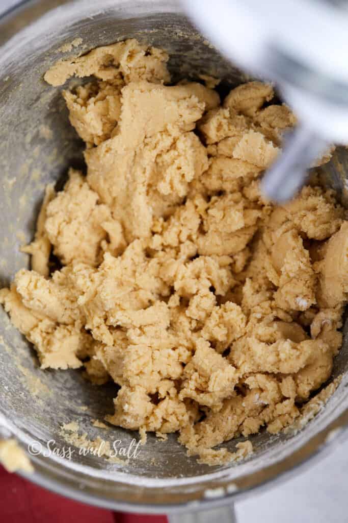 This image shows a close-up view of a bowl containing freshly mixed cookie dough. The dough appears soft and textured, indicative of being just combined to the right consistency for shaping into cookies. The mixing bowl is metallic, and part of a mixing spoon or spatula can be seen in the upper right corner, partially blurred, suggesting the action of mixing.