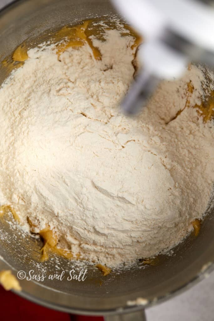 The image shows a top-down view of a stainless steel mixing bowl where a mound of flour has been added on top of a previously creamed mixture of butter and sugar. The mixing paddle attachment is in the midst of combining these ingredients, evident from the flour starting to integrate with the mixture beneath. The creamy yellow of the butter and sugar blend contrasts with the white flour, indicating the initial stage of combining wet and dry ingredients in a baking process.