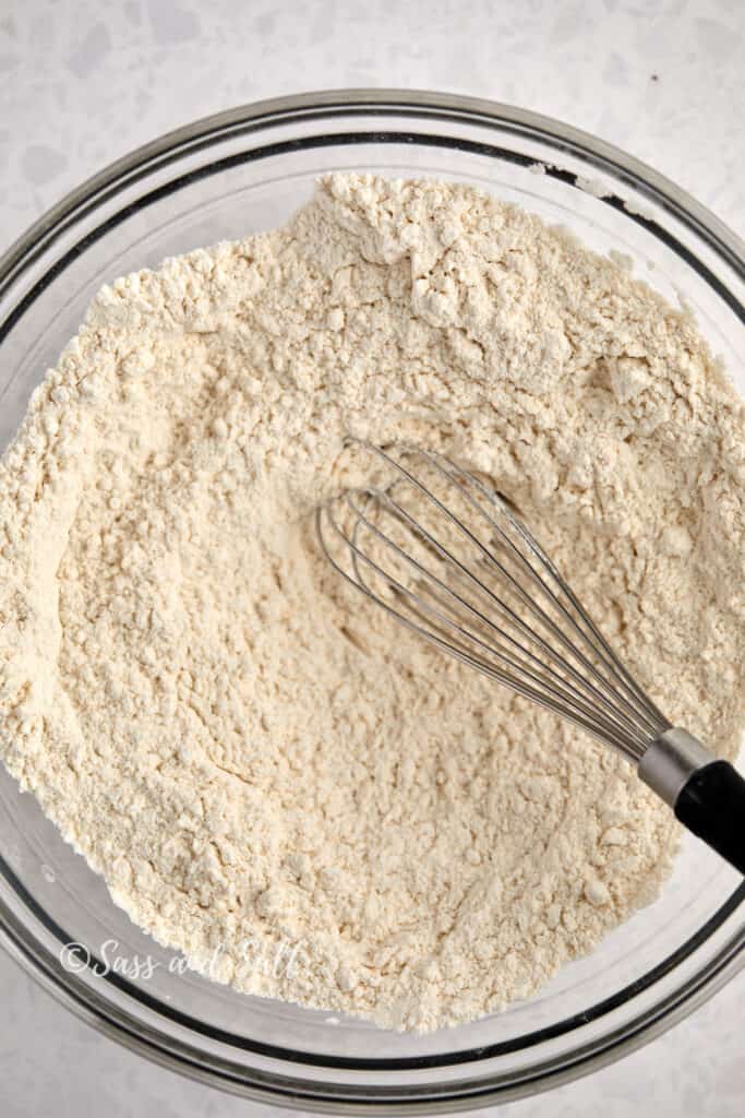 The image shows a clear glass mixing bowl filled with a dry mixture of flour, baking soda, baking powder, and salt. A whisk is in the bowl, indicating that these ingredients are being combined before adding to a wet mixture.
