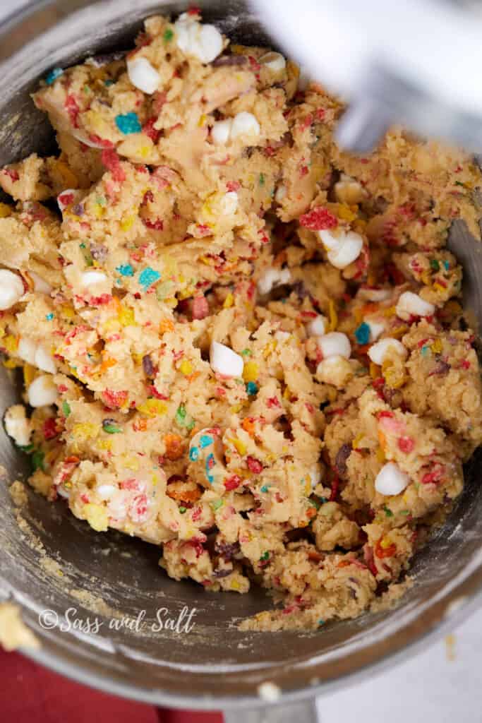 The image shows a close-up of a cookie dough mixture in a stainless steel mixing bowl, where colorful Fruity Pebbles cereal and white mini marshmallows have been thoroughly mixed into the dough. The mixture is vibrant with the specks of cereal adding a rainbow of colors throughout the golden brown dough.