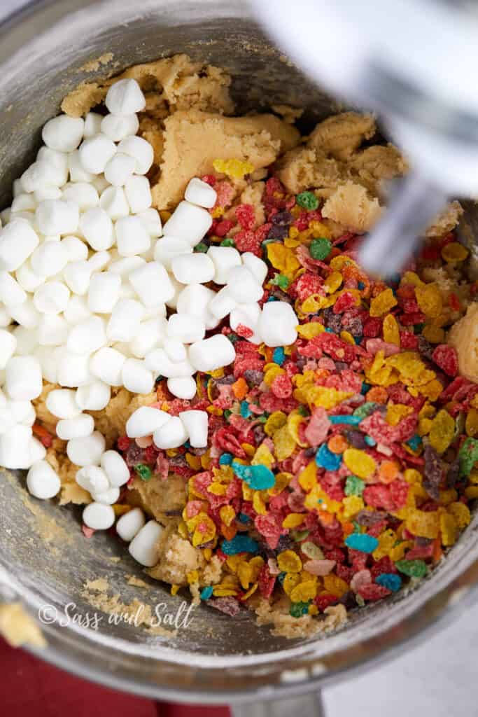 The image shows a stainless steel mixing bowl where a cookie dough mixture containing creamed butter and sugar is being combined with vibrant Fruity Pebbles cereal and white mini marshmallows. The colorful cereal and marshmallows are scattered across the top of the dough, ready to be mixed in, adding a pop of color and texture to the batch.