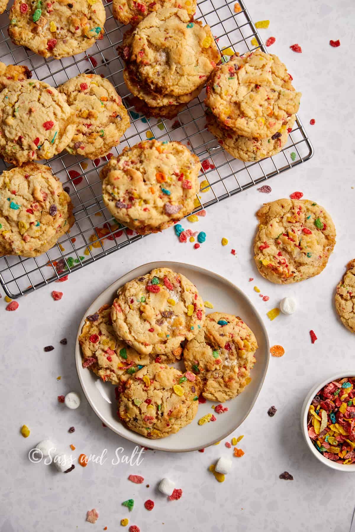 The image showcases a delightful array of Fruity Pebbles cookies presented on a cooling rack and a plate, with the scene set on a light, textured surface. The cookies are generously studded with vibrant pieces of Fruity Pebbles cereal, giving them a playful and colorful appearance. A scattering of cereal and a few marshmallows are artfully placed around the plate, enhancing the visual appeal of the homemade treats.
