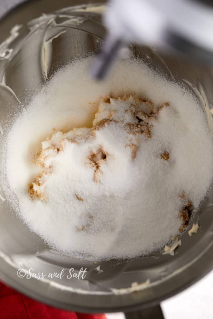 The image shows a mixing bowl where granulated sugar and brown sugar are being added to creamed butter. The white granulated sugar is piled high, with the golden brown sugar beneath it, creating a contrasting mound against the pale, whipped butter.