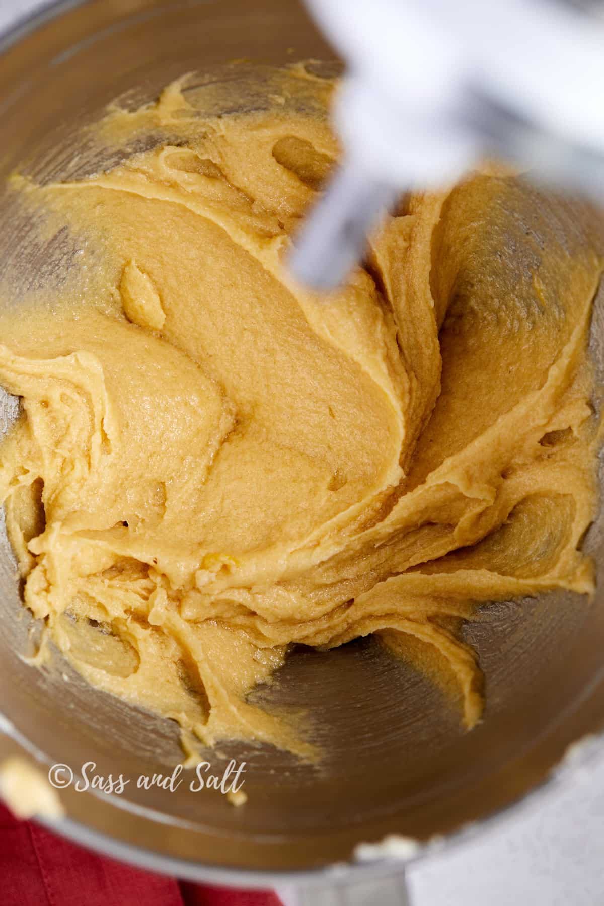 The image shows a close-up view of a mixing bowl where butter and sugar have been creamed together, forming a light, fluffy, and pale yellow mixture. The mixing paddle attachment, in motion, is creating swirls in the mixture, indicating the process of creaming is well underway. The sides of the bowl have been scraped clean, ensuring even mixing.