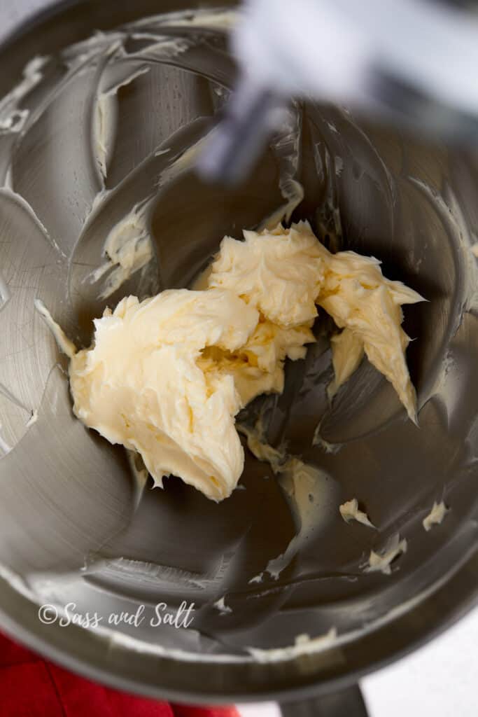 The image captures a close-up view inside a stainless steel mixer bowl, where unsalted butter has been partially creamed, showing a soft, whipped texture. The mixing paddle attachment is visible at the top, moving through the butter, which clings to the sides of the bowl indicating the beginning stages of creaming.