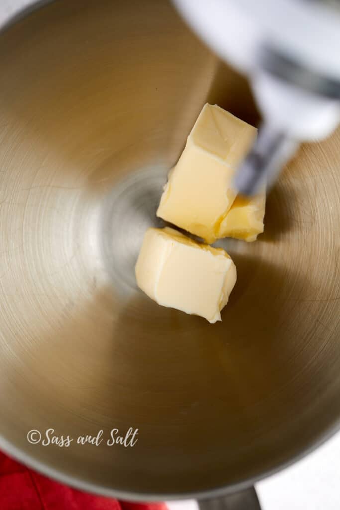 The image displays a close-up view of a stainless steel mixing bowl from a stand mixer, with two large chunks of unsalted butter at the bottom. The butter appears soft, indicating it's at room temperature, which is ideal for creaming in baking. In the background, the mixer's attachment is seen hovering over the butter, ready to start the creaming process.