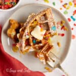A stack of golden waffles topped with a pat of butter, sprinkled with confectioners' sugar and colorful cereal, served on a white plate with a fork ready for a sweet and whimsical breakfast indulgence.