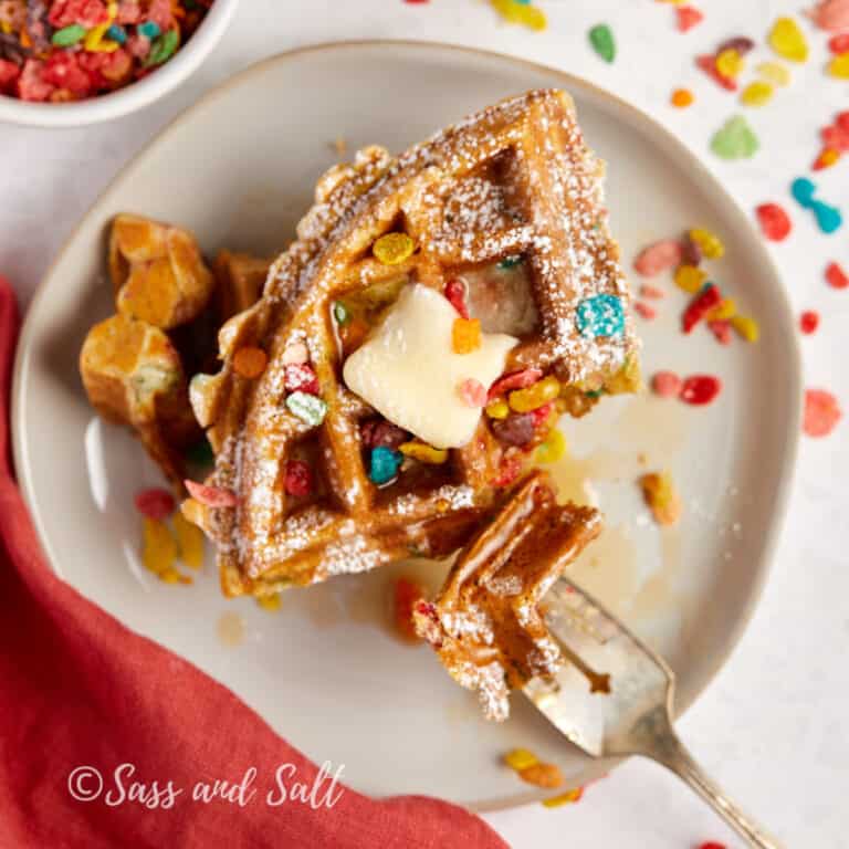 A stack of golden waffles topped with a pat of butter, sprinkled with confectioners' sugar and colorful cereal, served on a white plate with a fork ready for a sweet and whimsical breakfast indulgence.