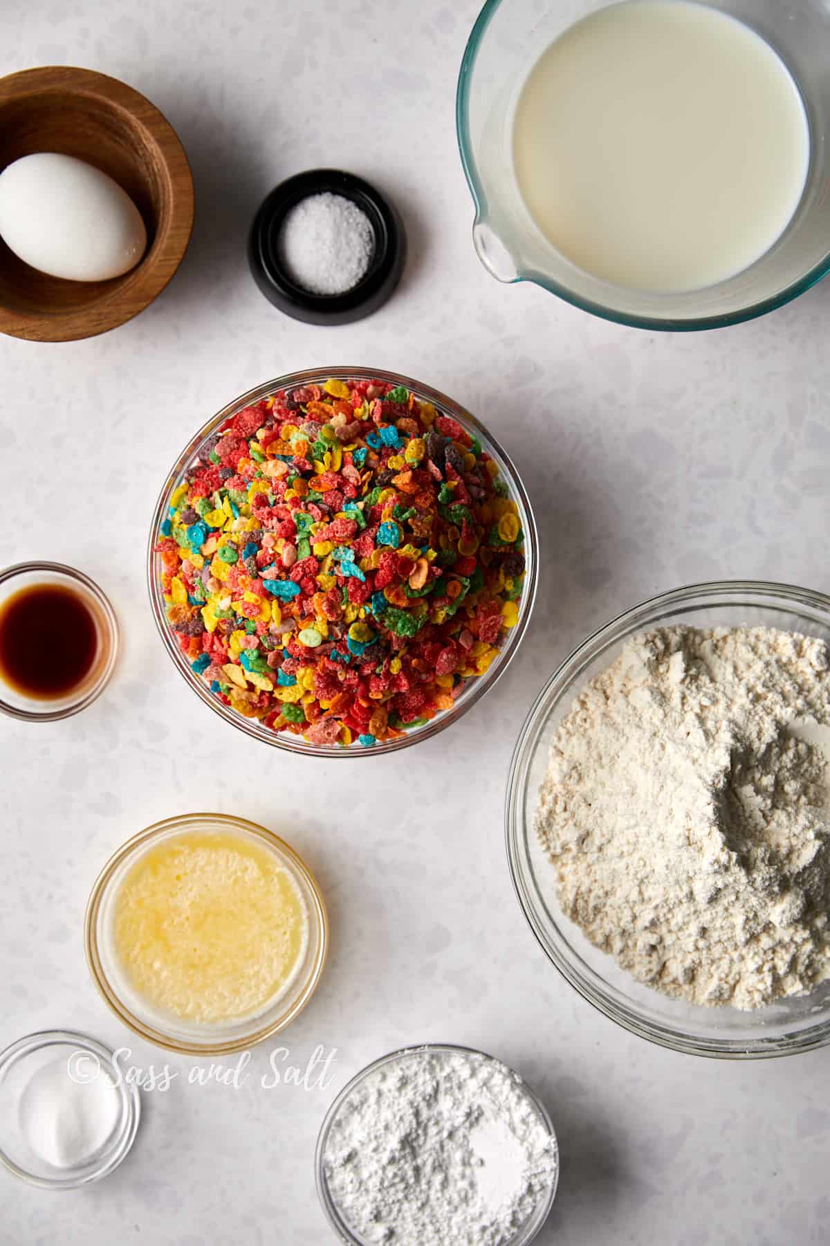Ingredients for baking arranged on a table: Fruity Pebbles cereal in a bowl, eggs, salt, milk, vanilla extract, melted butter, flour, and confectioner's sugar, with a playful "sass and salt" watermark.