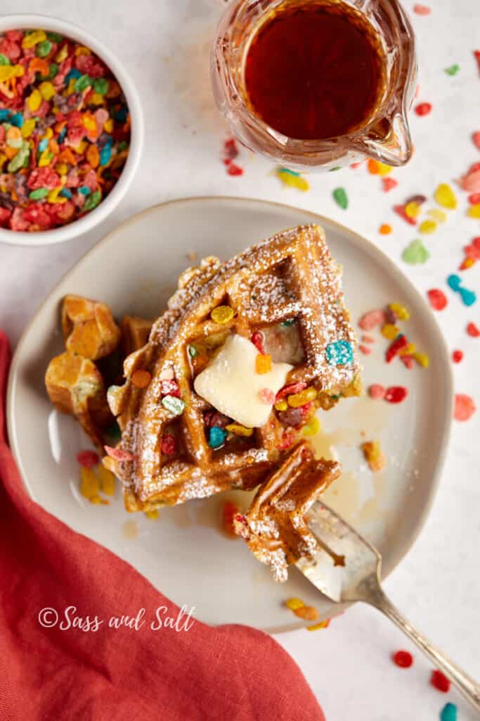 Plate of waffles topped with colorful cereal, a dollop of butter, and syrup being poured over, with a playful scattering of cereal pieces around the plate.