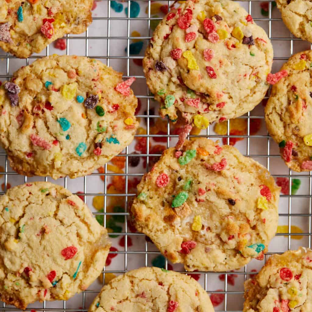 The image shows freshly baked cookies cooling on a wire rack. These treats are packed with colorful Fruity Pebbles cereal pieces that add a vibrant contrast to the golden-brown hue of the cookies.