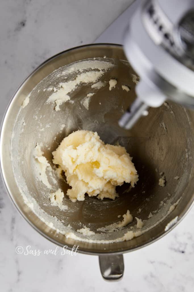 Creamed butter and sugar mixture in a stainless steel mixing bowl with a mixer above it, set on a marble countertop.
