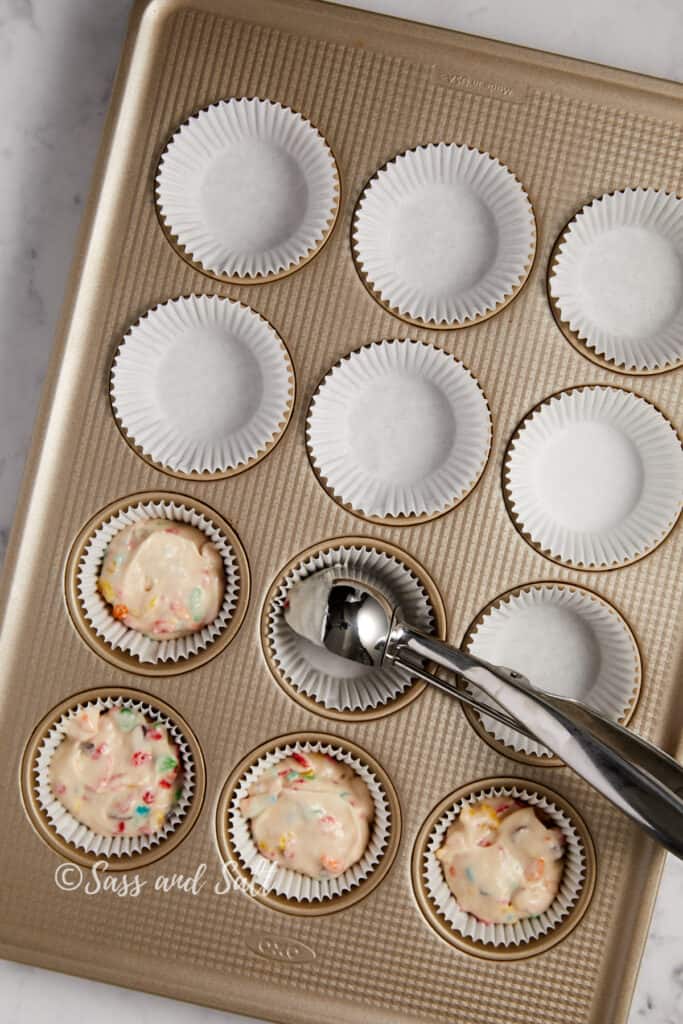 A gold cupcake tray with white paper liners, some filled with speckled cupcake batter. A cookie scoop rests in the tray, used for portioning, all under a "Sass and Salt" watermark.