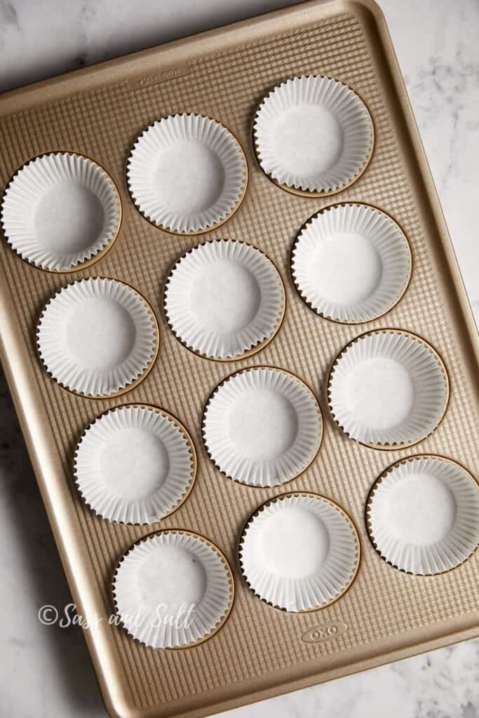 A gold-colored cupcake pan with white paper liners placed in each mold, set on a marble countertop.