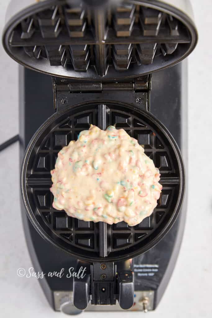 
A scoop of colorful Fruity Pebbles waffle batter is placed in the center of an open, preheated waffle iron, ready to be cooked to a perfect golden crisp. The image showcases the beginning of the waffle-making process and is marked with "Sass and Salt" branding.