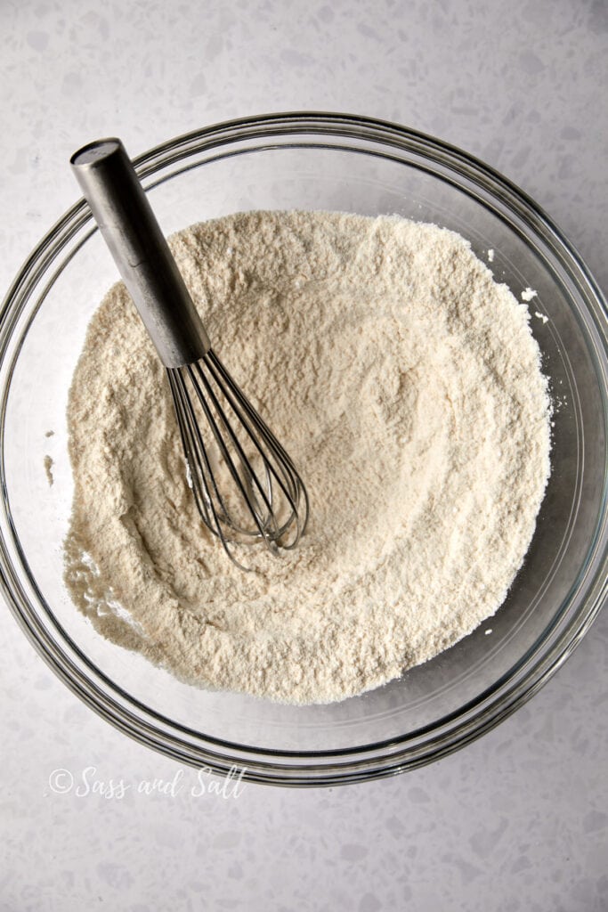 A clear glass bowl filled with flour and other dry ingredients mixed together.  A whisk is sitting on the side of the bowl.